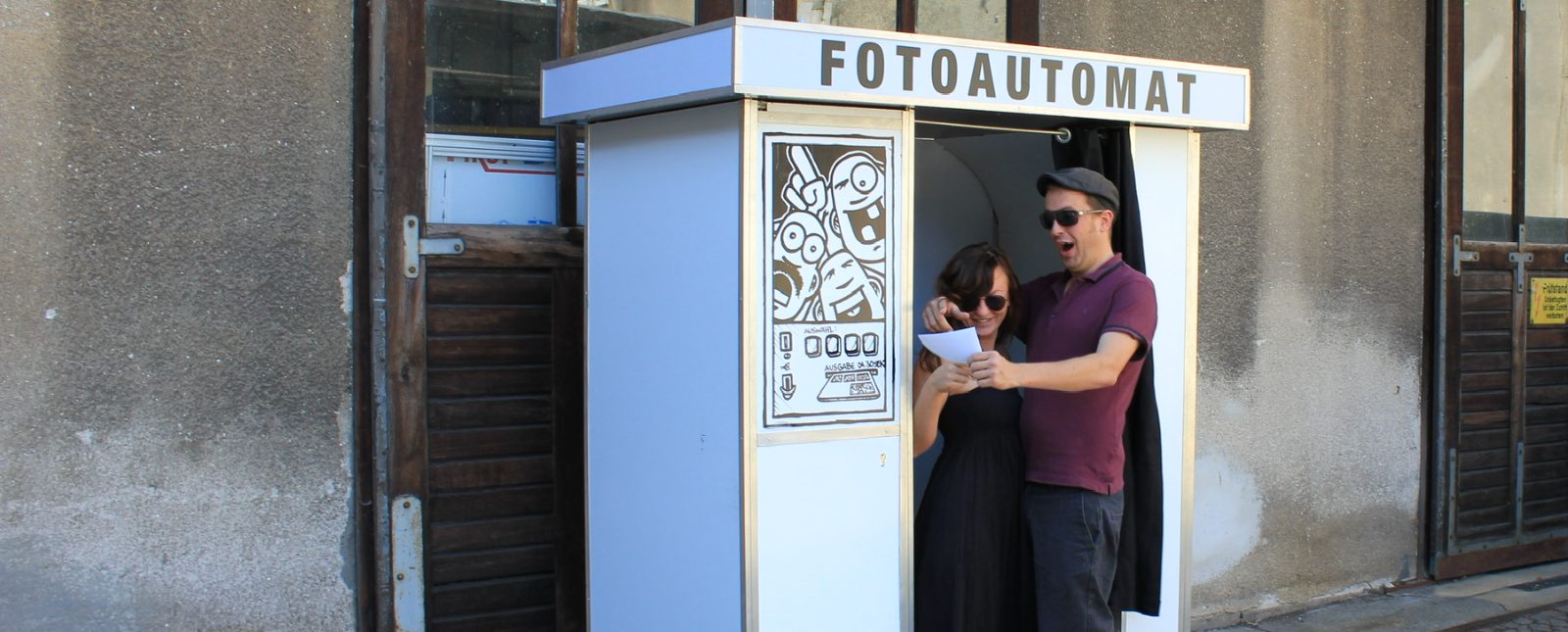 Lachendes Paar zeigt sich das frisch gedruckte Foto vor dem klassischen Fotoautomaten von Flash-U