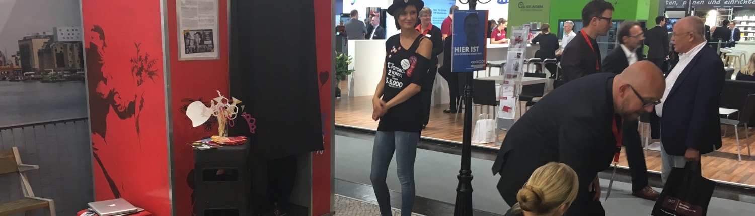 fu-messen-hero-photoautomat Besucherin lächelt neben dem roten Fotoautomaten von Flash-U auf einem Messestand